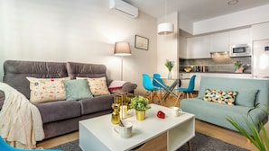 Een moderne woonkamer met een grijze bank, een witte salontafel en een blauwe fauteuil. Op de achtergrond is een keuken zichtbaar met witte kastjes en een magnetron.
