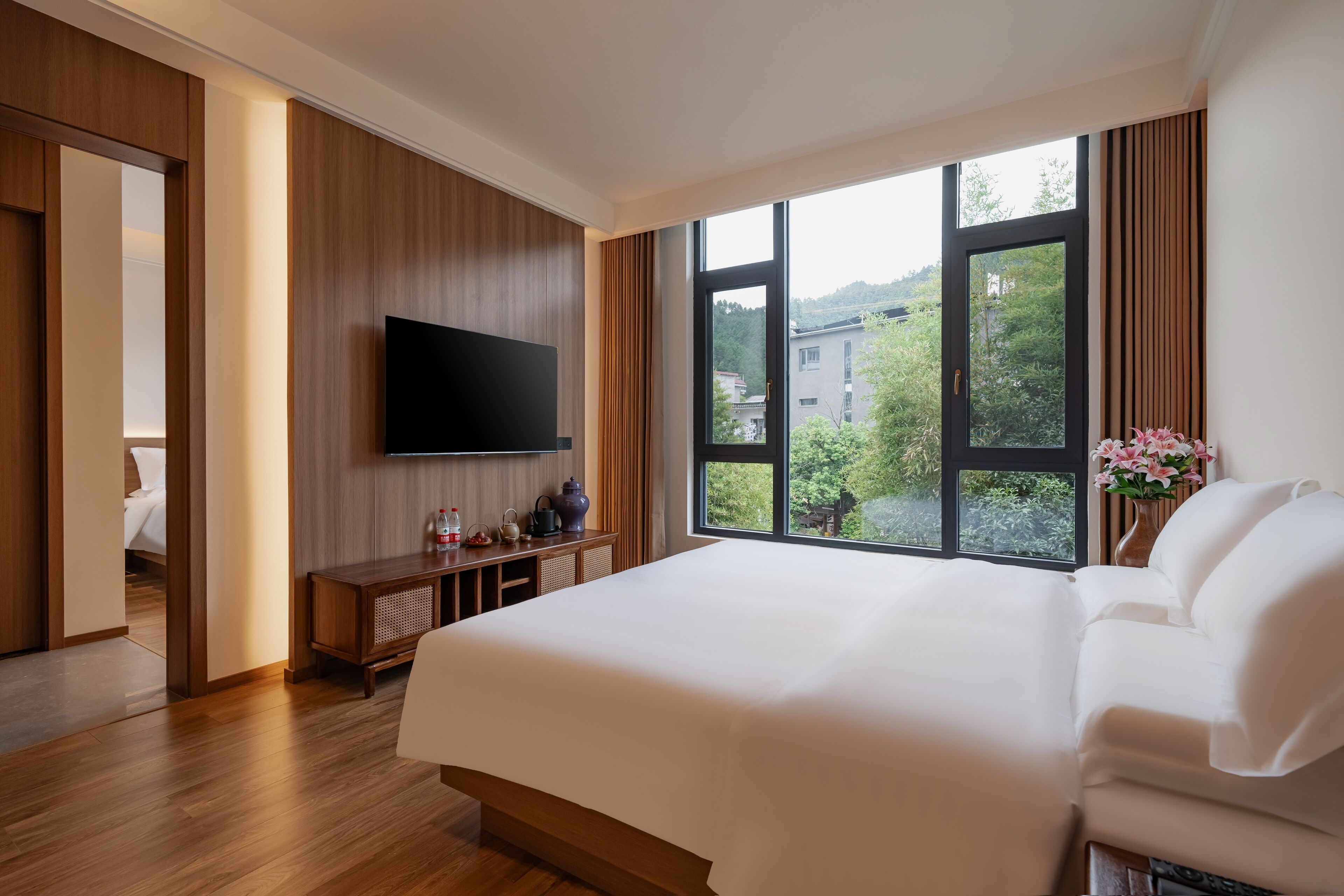 A modern hotel room with a large bed, a flat-screen TV mounted on the wall, a wooden console table, and a large window offering a view of the outdoors.