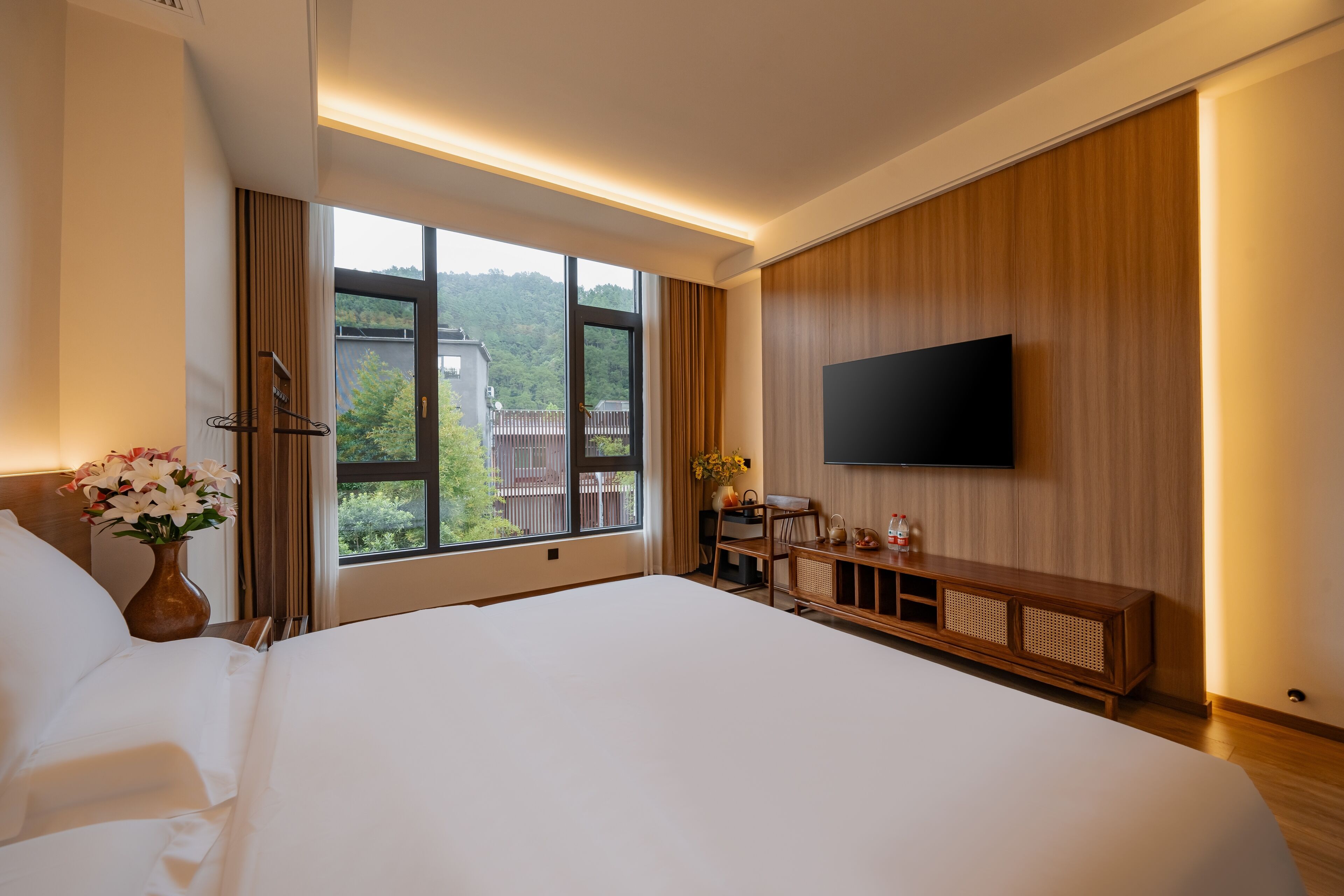 A hotel room with a large bed, a TV mounted on the wall, a wooden cabinet, and a window with a view of greenery.