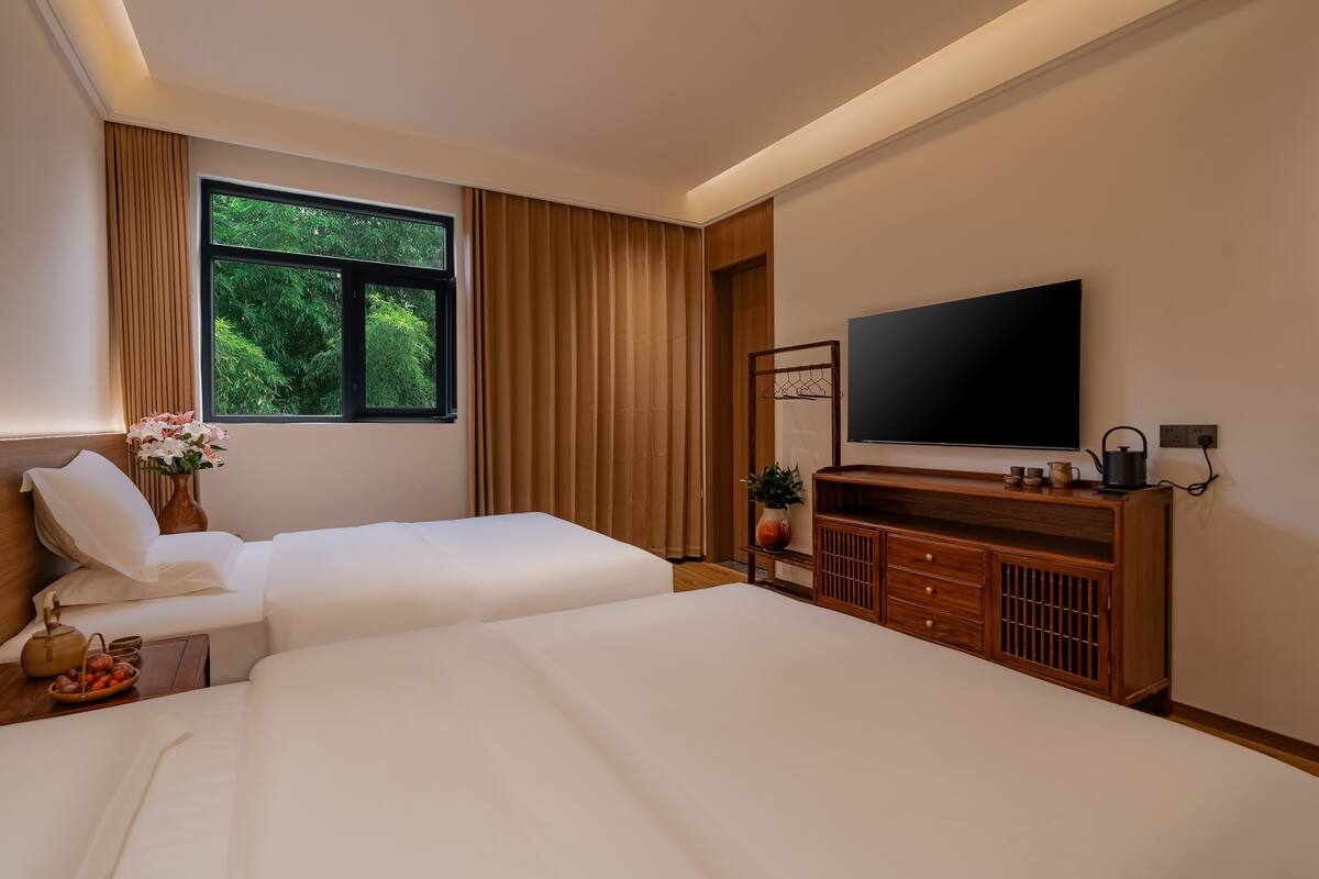 A hotel room with two beds, a TV, a wooden dresser, and a window with a view of greenery.