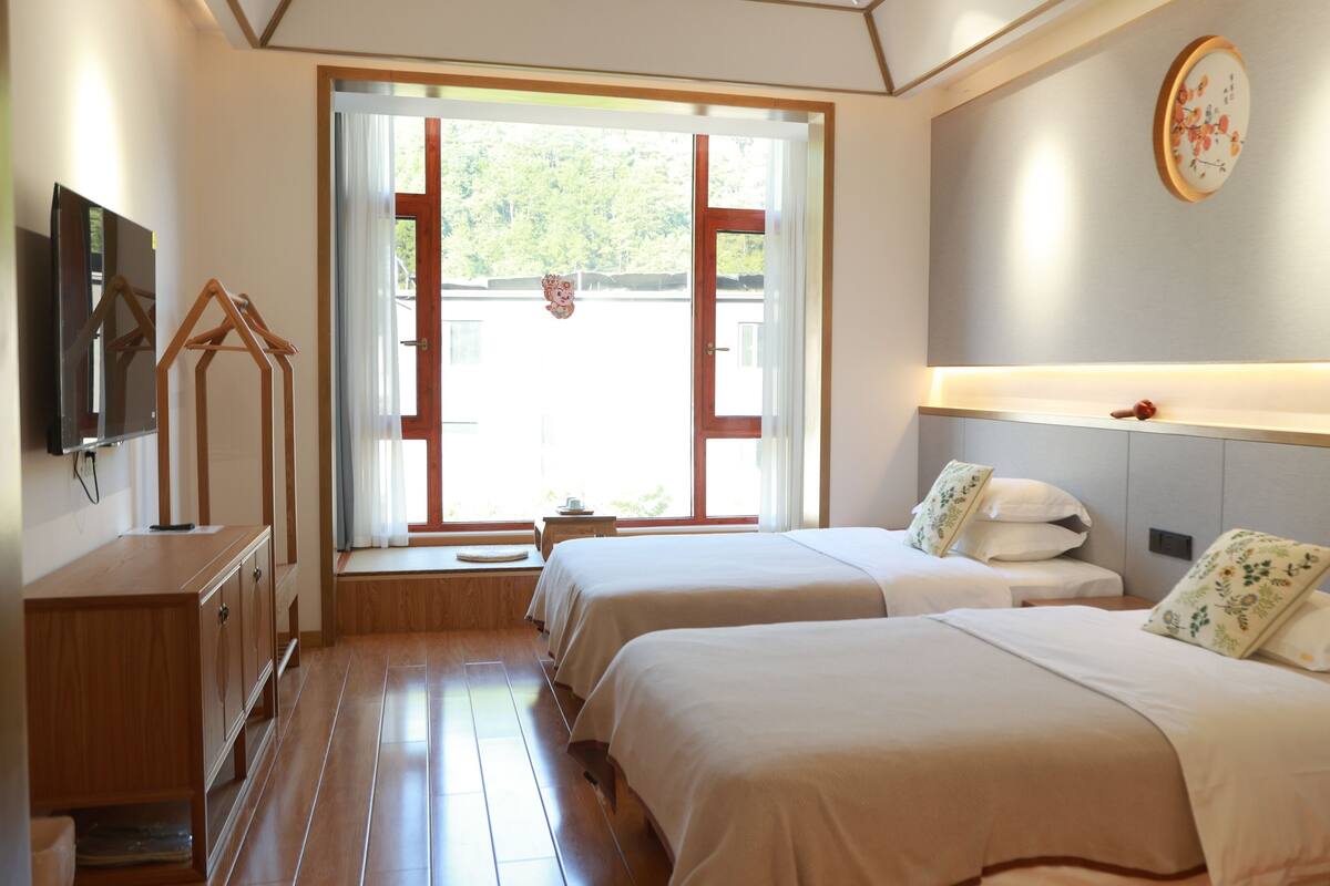 A hotel room with two beds, a TV, a wooden desk, and a window with a view of greenery.