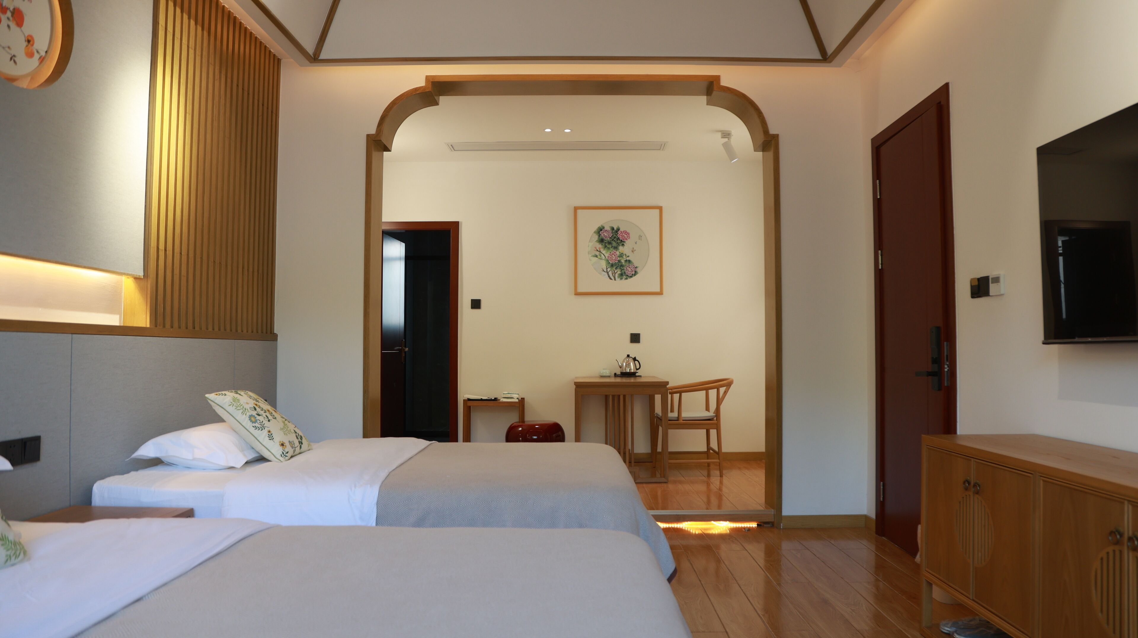 A hotel room with two beds, a wooden wardrobe, a desk with a chair, and a television mounted on the wall.