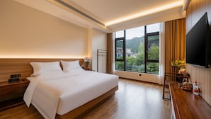 Une chambre d’hôtel moderne dotée d’un grand lit, de tables de chevet, d’un téléviseur à écran plat et d’une fenêtre donnant sur un paysage verdoyant et des bâtiments.