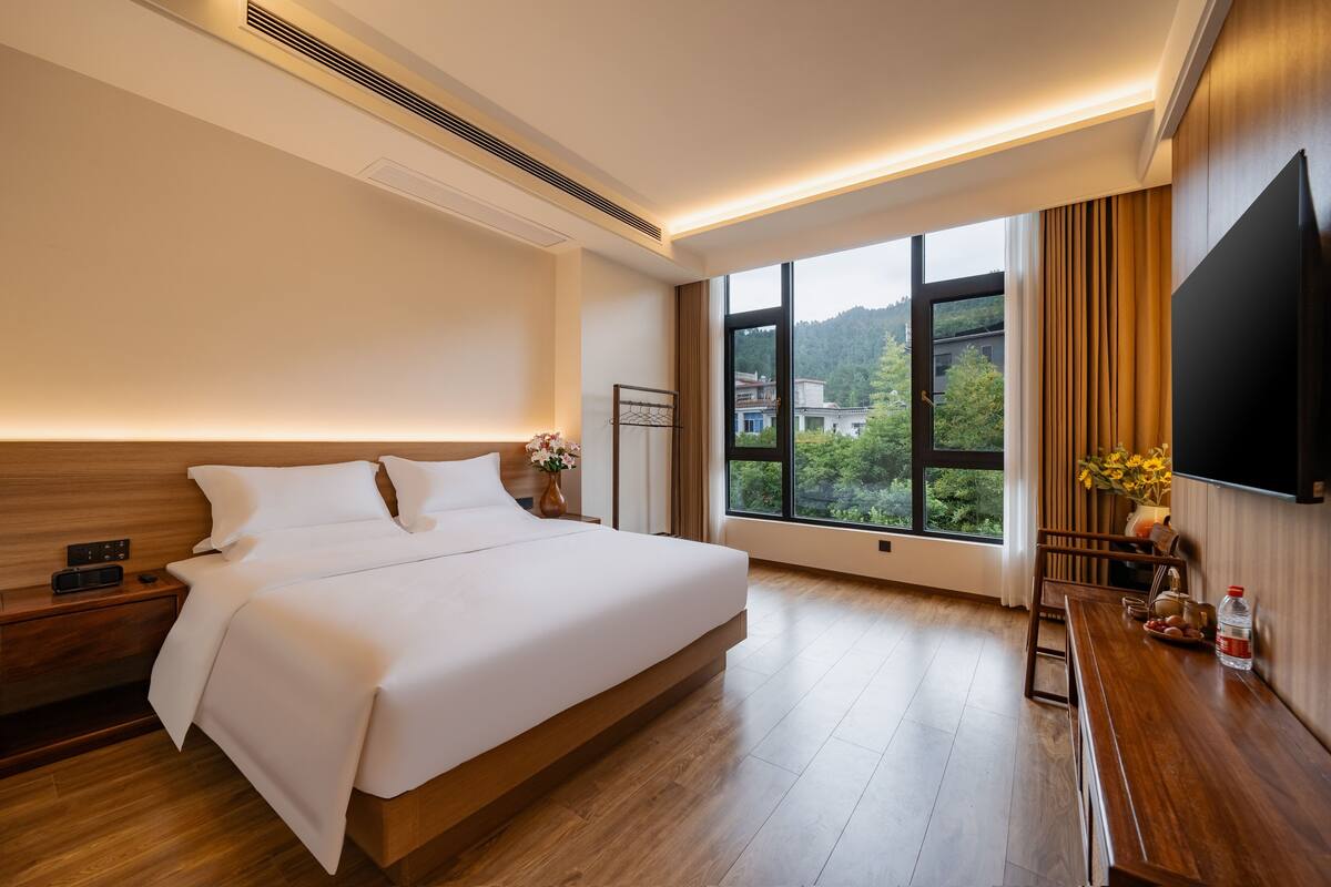 A modern hotel room with a large bed, bedside tables, a flat-screen TV, and a window with a view of greenery and buildings.