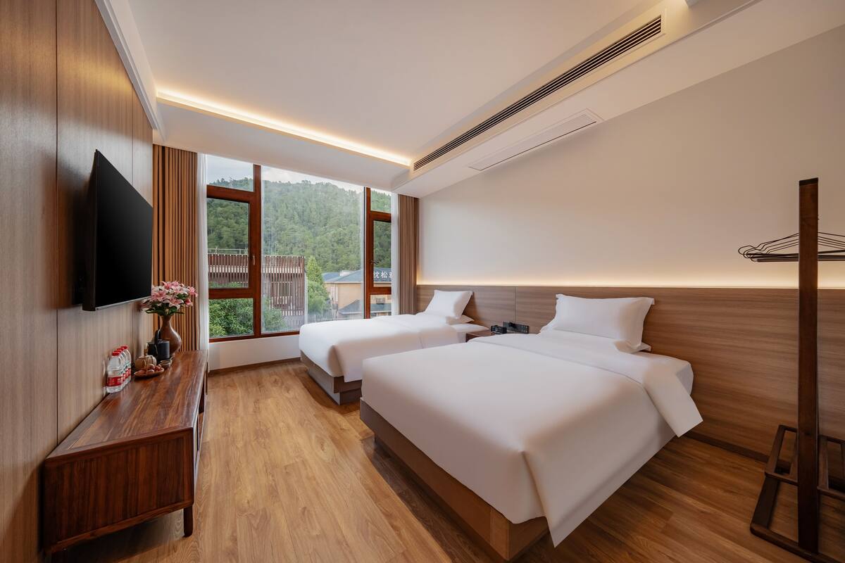 A hotel room with two beds, a wooden bench, a flat-screen TV, and a large window offering a view of a forested area.