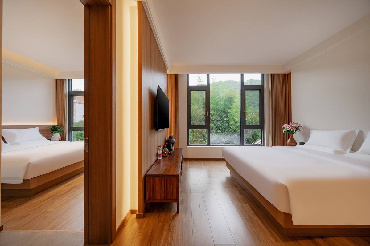 A hotel room with two beds, a wooden floor, a TV on the wall, and large windows with a view of greenery outside.