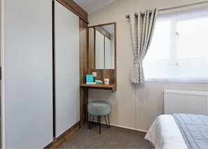 Ein Schlafzimmer mit Bett, einem Fenster mit Vorhängen, einem Spiegel, einem kleinen Hocker und einem Regal mit Pflegeprodukten.