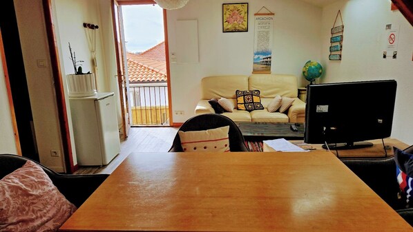 A cozy living room with a wooden dining table, a sofa with patterned cushions, a television, and a small refrigerator.