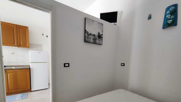 A bedroom with a bed, a small kitchenette with wooden cabinets, a white refrigerator, and a wall-mounted TV. - Studio apartment in downtown (Realmonte)