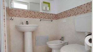 A bathroom with a pedestal sink, a bidet, and a toilet. There is a mirror above the sink and a small shelf with toiletries. - Studio apartment in downtown (Realmonte)