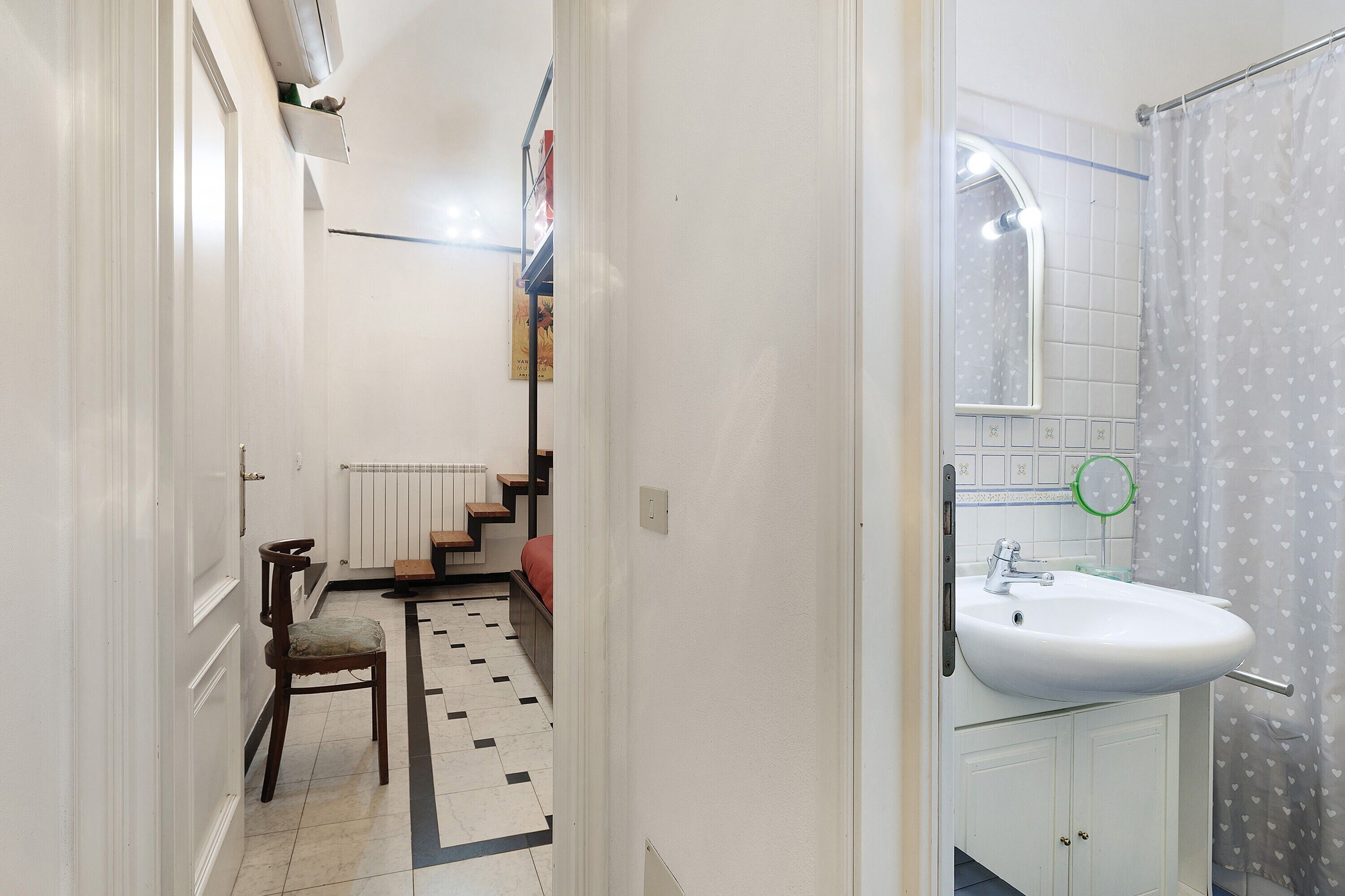 Ein schmaler Flur, der zu einem Badezimmer führt, mit einem weißen Waschbecken, einer Dusche mit einem Herzmuster-Vorhang und einem Stuhl.