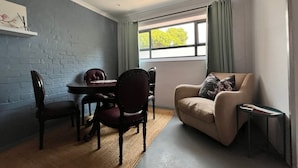Ein Zimmer mit Esstisch und Stühlen, einer Couch, einem Fenster mit Vorhängen und einem kleinen Tisch mit einem Buch.