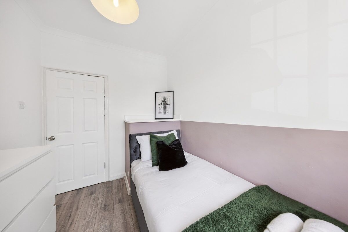 A bedroom with a bed, a white door, a framed picture on the wall, and a light fixture.
