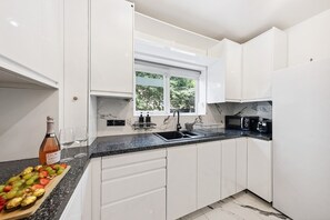 A modern kitchen with white cabinets, a black countertop, a sink, and a window. - Stylish and Spacious 3 BR Apartment + Cozy Garden (Greater London)