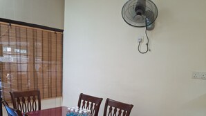 A dining area with a table set for four, surrounded by wooden chairs. A fan is mounted on the wall above the table, and a window with a bamboo blind is visible in the background. - Spacious 4-bedroom house in gorgeous Subang Jaya with AC (Subang Jaya)