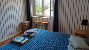 A bedroom with striped wallpaper, a bed with a blue quilt, a wooden bench, a radiator, and a window with a view of greenery. - Unique Large Home in Estoril with Pool - Perfect for Families (Estoril)