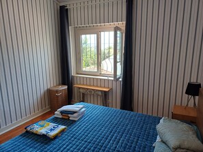 A bedroom with striped wallpaper, a bed with a blue quilt, a wooden bench, a radiator, and a window with a view of greenery. - Serene 3-bedroom house in Estoril perfect for relaxing getaways (Estoril)