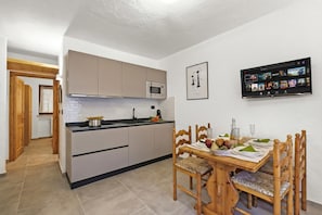A modern kitchen with a built-in microwave, a dining table set with plates and glasses, and a flat-screen TV mounted on the wall displaying a movie. - Rochebrune Apartments (Claviere)