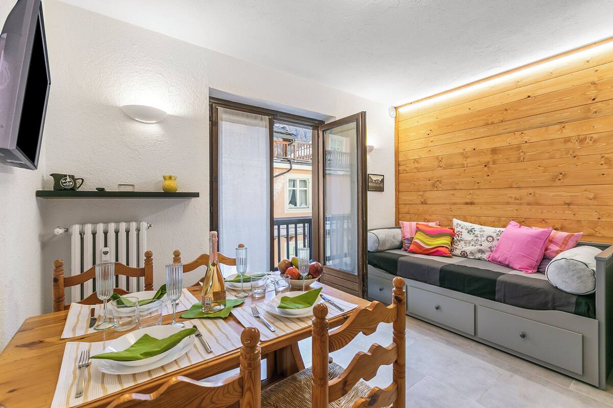 A dining area with a table set for two, a sofa with cushions, a television, and a view through a glass door.