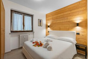 Una cama bien hecha con toallas y un libro, una ventana con cortinas, un radiador y paneles de madera en la pared.
