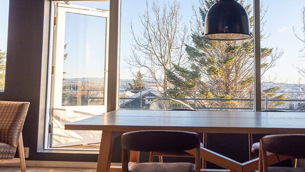 Uma área de jantar moderna com uma mesa grande e cadeiras, vista para árvores e edifícios através de uma ampla janela, e uma luminária pendente sobre a mesa.