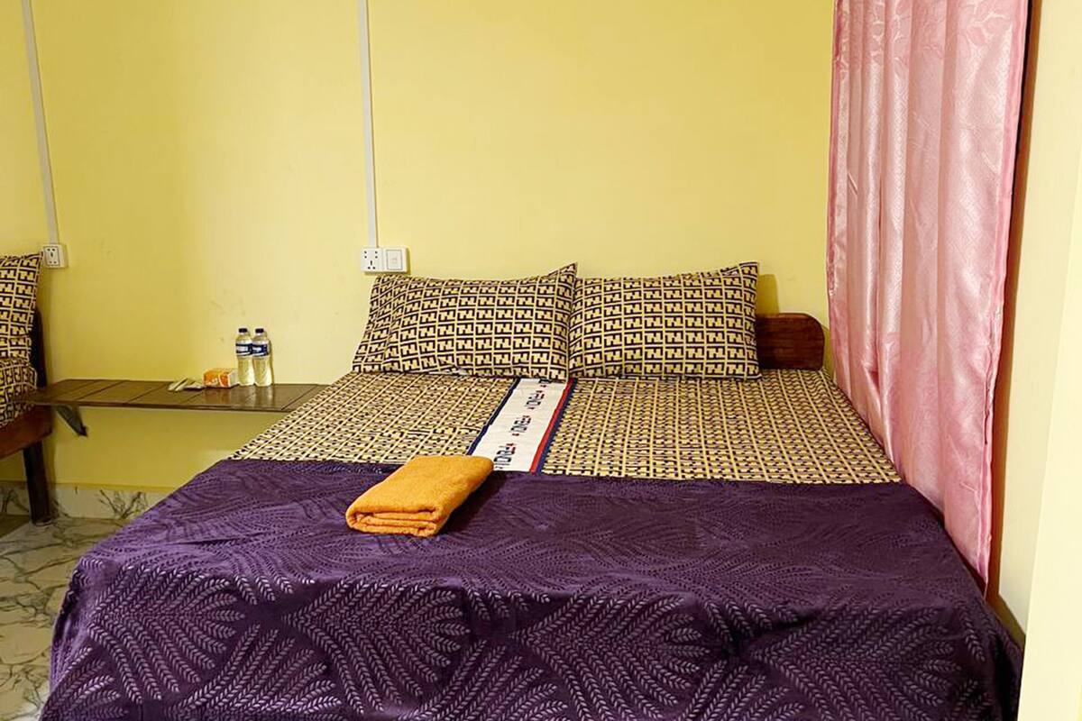 A hotel room with a bed, pillows, a blanket, a curtain, and a small table with bottles.