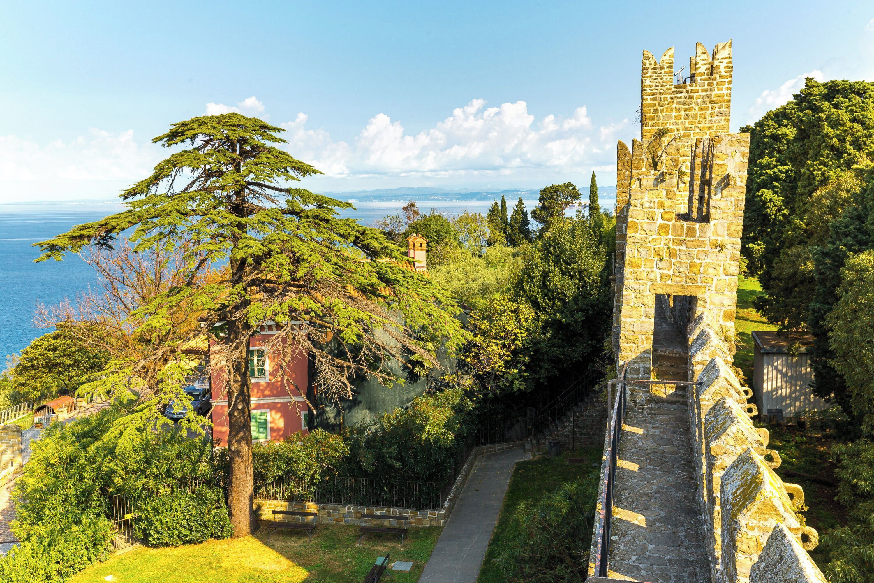 Un château doté d’une tour en pierre et d’un sentier menant à celui-ci, entouré d’une végétation luxuriante et offrant une vue sur la mer.