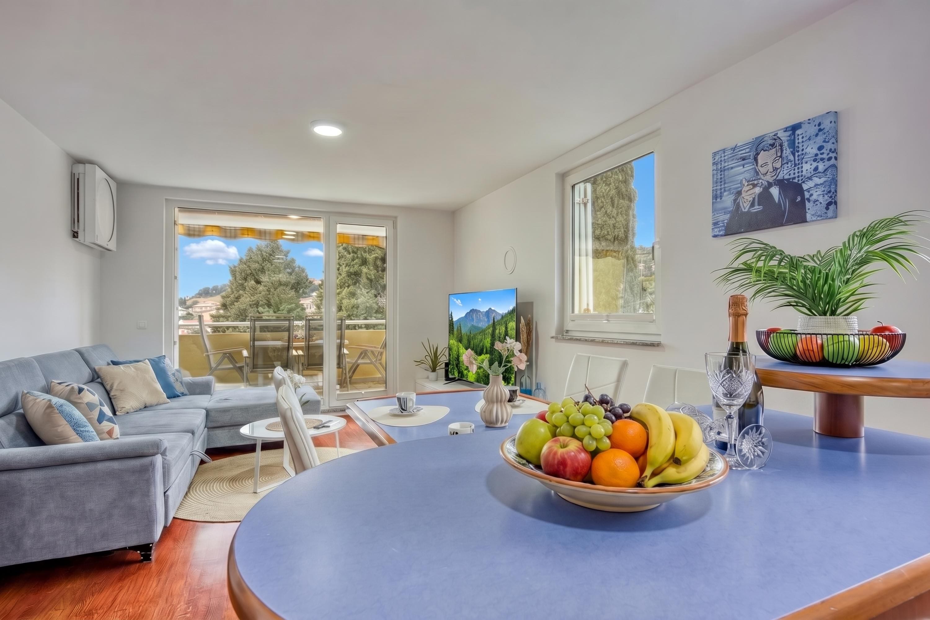 A modern living room with a grey sofa, a dining table with fruit and a bottle, and a large window with a city view.