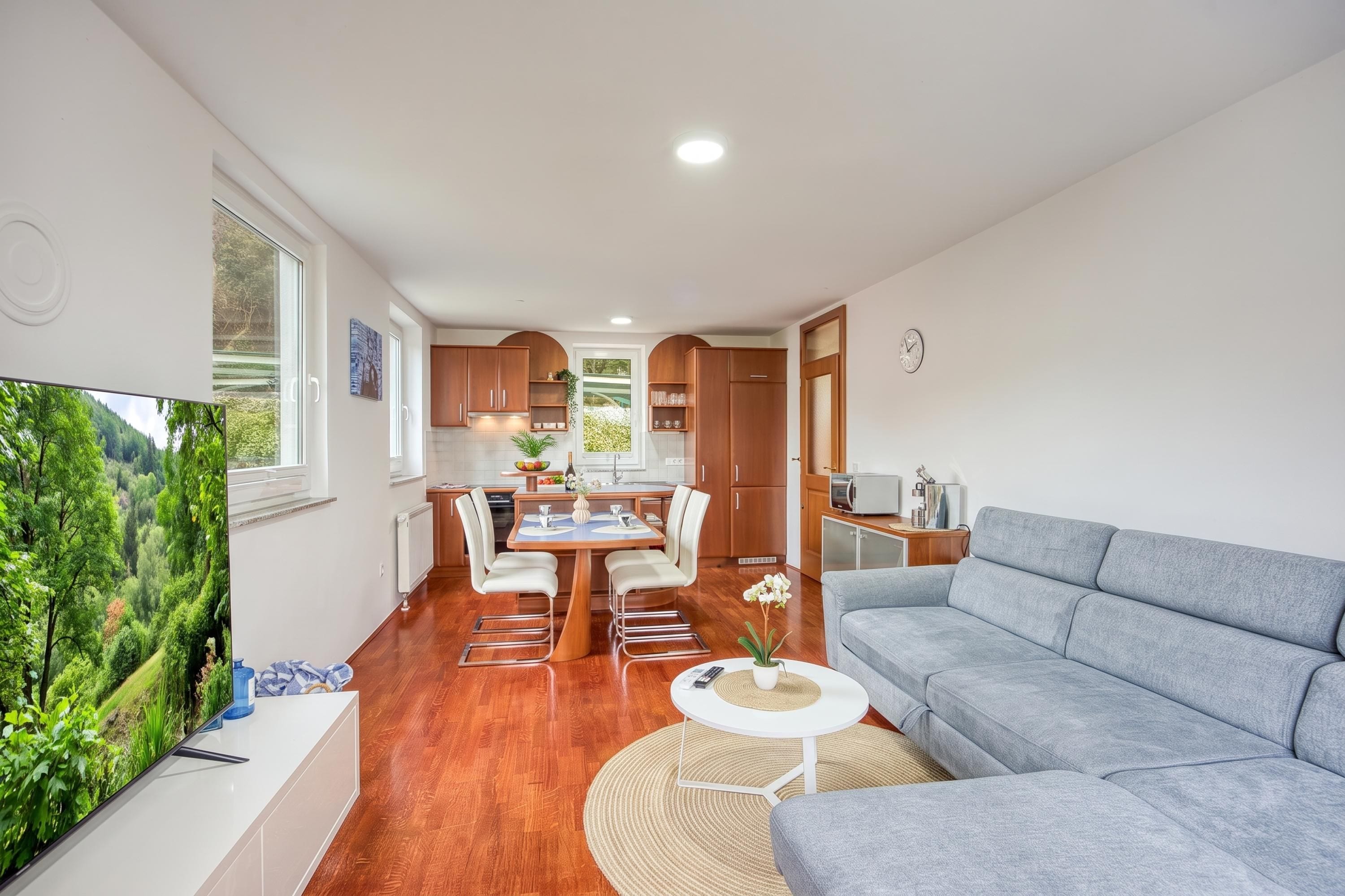 A modern living room with a grey sofa, a dining area with a table and chairs, and a kitchen with wooden cabinets.