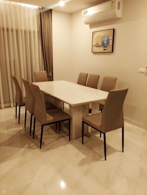 Un coin repas avec une table rectangulaire entourée de six chaises, une grande fenêtre avec des stores verticaux et un appareil de climatisation fixé au mur.
