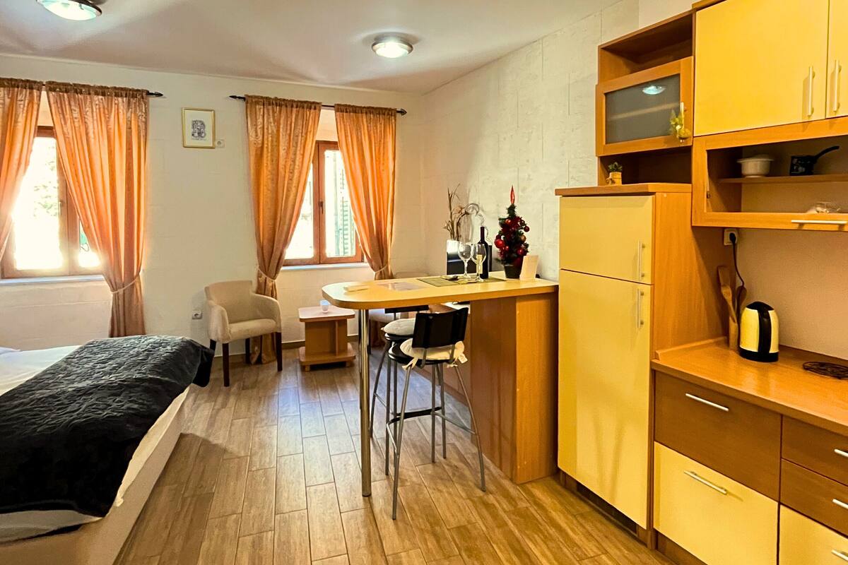 A compact room with a bed, a small dining table, a chair, and a kitchenette area.