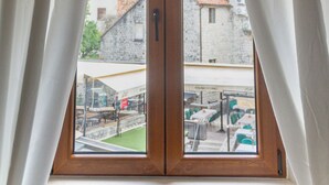 A window with white curtains, a view of buildings, and a green area outside. - Step to Kotor Bay Old Town (Kotor)