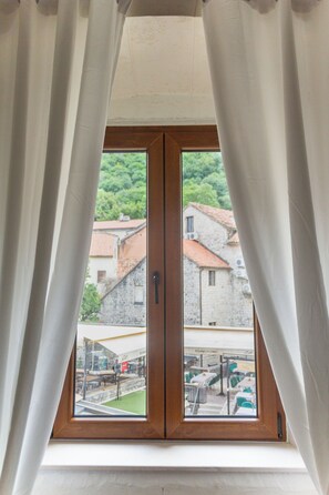 A window with white curtains, a view of buildings, and a green area outside. - Step to Kotor Bay Old Town (Kotor)
