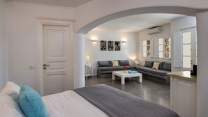 Ein modernes Schlafzimmer mit einem großen Bett, einem grauen Sofa, einem weißen Couchtisch und einem kleinen Tisch mit einer Lampe. Links ist eine weiße Tür, und ein weißer Durchgang führt zu einem Wohnbereich mit einem grauen Sofa und einer Steinwand.