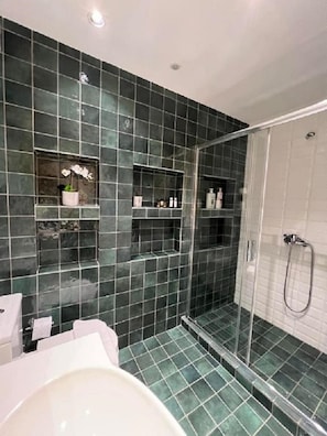 A modern bathroom with dark green tiles, a glass shower enclosure, and built-in shelves with toiletries. - Crown Syntagma Apartment (Athens)