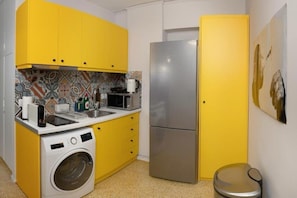A compact kitchen with yellow cabinets, a stainless steel refrigerator, a washing machine, and a sink. - Crown Syntagma Apartment (Athens)