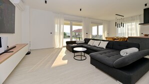 A modern living room with a grey sofa, a small round table, and a large window with curtains. - Villa Giovanni Zadar Villas (Razanac)