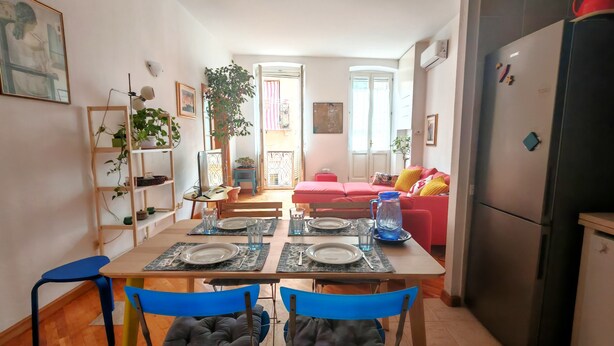Zona pranzo con tavolo apparecchiato per quattro, cucina con frigorifero e soggiorno con divano rosso.