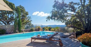 Um deck de madeira com duas espreguiçadeiras e uma piscina, cercado por árvores e um muro de pedra.