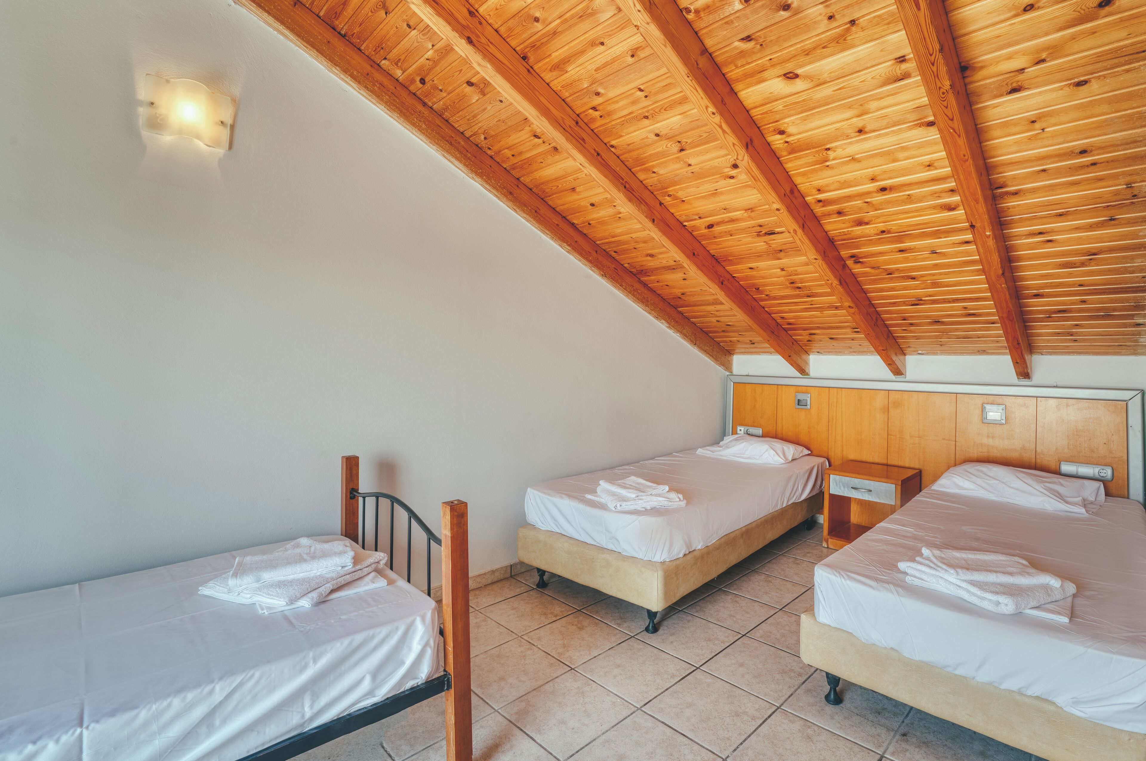 Une chambre avec deux lits simples, des poutres apparentes en bois au plafond, un luminaire fixé au mur et un sol carrelé.
