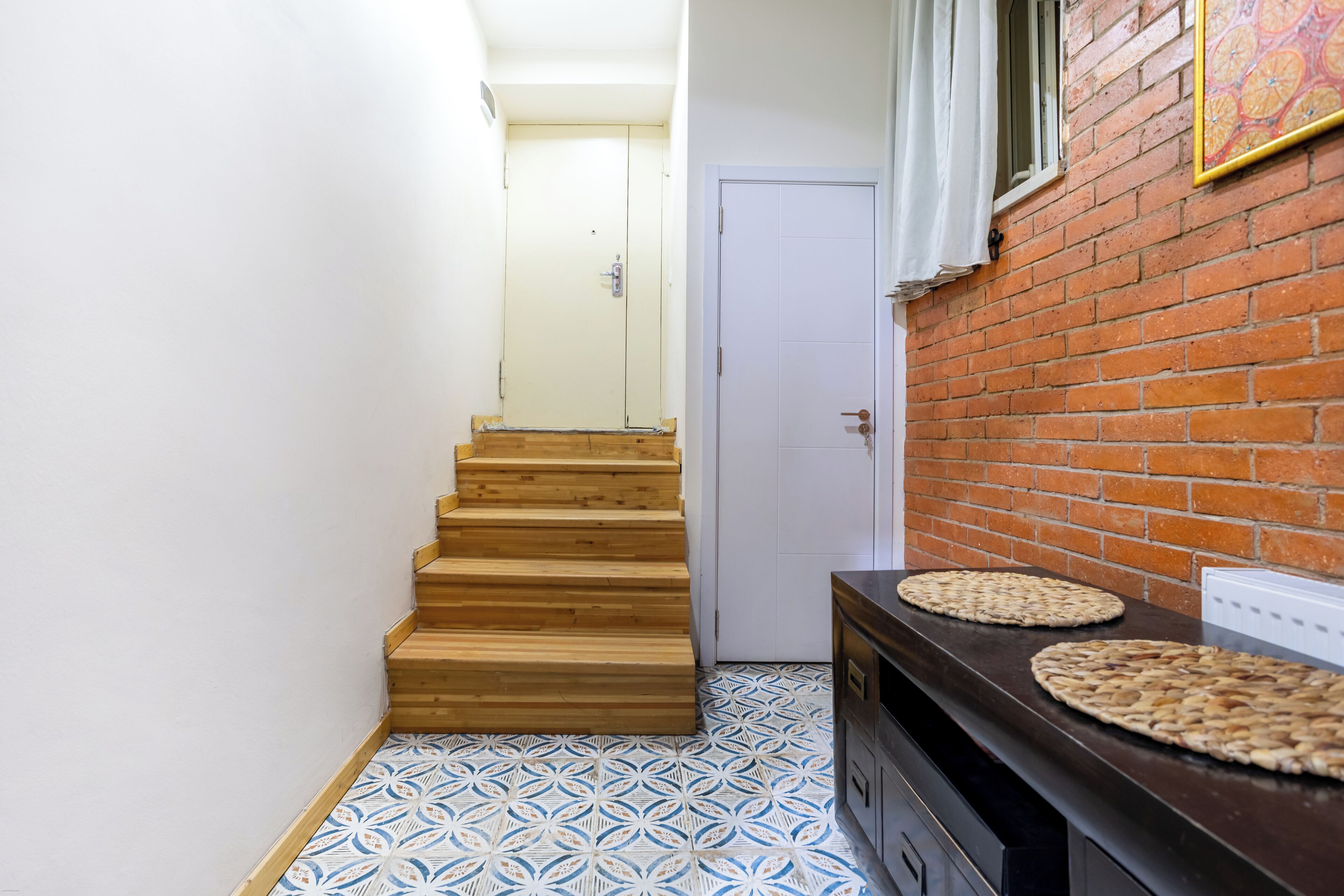 A narrow hallway with a wooden staircase leading to a door, a patterned floor, and a brick wall with a picture frame.