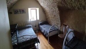Habitació petita i rústica amb sostre de pedra, dos llits de metall, un terra de fusta, una taula petita i una finestra.