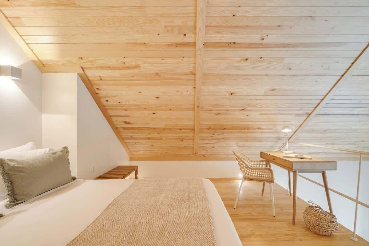 A modern bedroom with a sloped ceiling, a bed with a white mattress and a beige throw, a wooden desk with a chair, and a small table lamp.