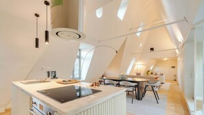 Cozinha moderna com ilha central, área de refeições com mesa redonda e quarto em mezanino com teto inclinado.