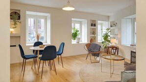 En rummelig opholdsstue med spisebord, stole, et rundt sofabord og en sofa. Store vinduer giver naturligt lys og udsigt til omgivelserne.