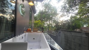 Un baño exterior moderno con una bañera grande, un estante de madera con frutas y una botella de vino, y vistas a árboles y un muro de ladrillo.