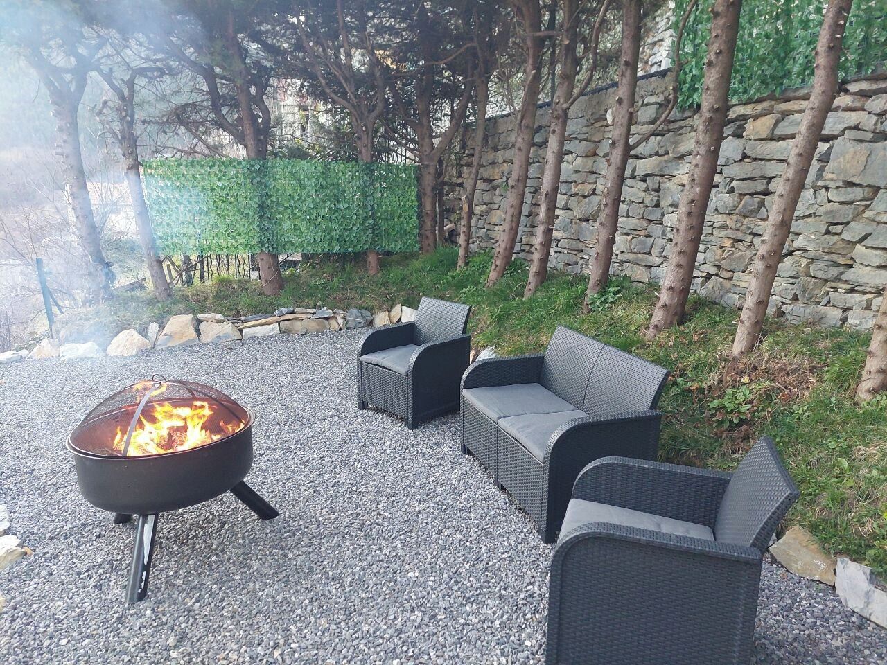 Een vuurkorf met twee rieten banken in een tuin met een stenen muur en bomen op de achtergrond.