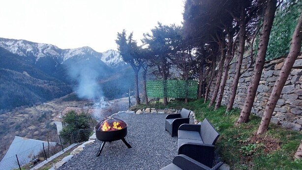 Een terras buiten met een vuurkorf, omringd door stenen muren en bomen, met uitzicht op een bergachtig landschap.