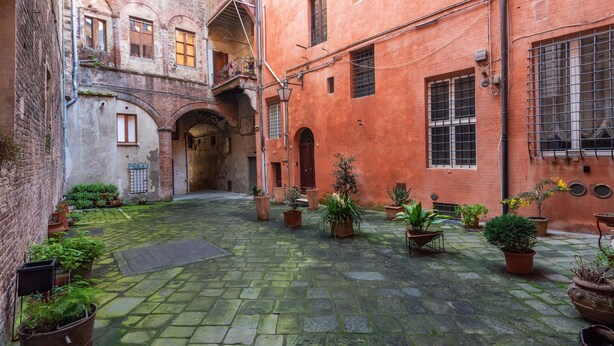 A courtyard with stone paving, potted plants, and a brick building with arched doorways. - Campo Apartment in the Center of Siena (Siena)