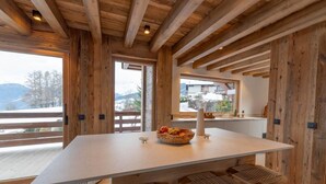 Interior em madeira com uma mesa grande e banquetas, uma janela com vista para montanhas e uma cesta de frutas sobre a mesa.